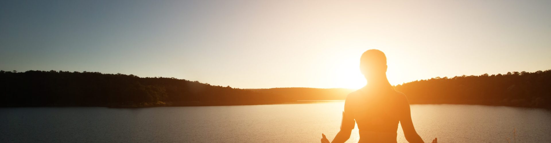 Silhouette of healthy woman is practicing yoga at mountain lake during sunset.